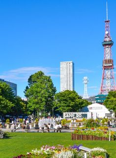 a park with lots of flowers and people walking around it in front of a tall tower