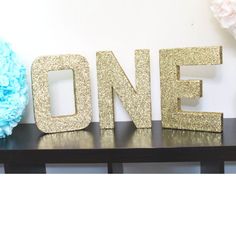 the word one is spelled with gold glitter letters on a black shelf next to blue flowers