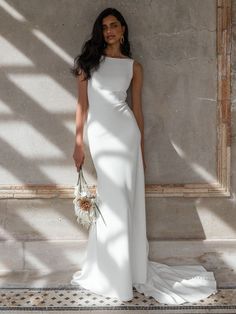 a woman standing in front of a wall wearing a white dress and holding a bouquet