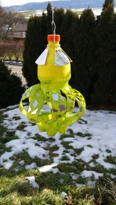 a yellow bird feeder hanging from a tree in the snow covered yard with grass and bushes