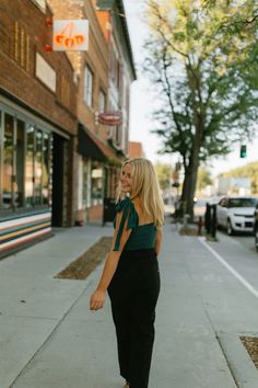 a woman standing on the sidewalk in front of a building wearing a green top and black pants