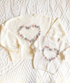 three baby sweaters are laid out on a white quilted bed with embroidered hearts