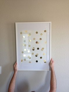 a person holding up a white framed artwork with gold dots on it's side