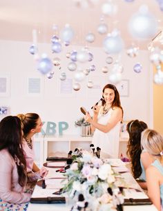 a woman standing in front of a table holding a microphone and talking to other women