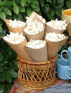 there is a basket full of popcorn on the table next to some cups and saucers