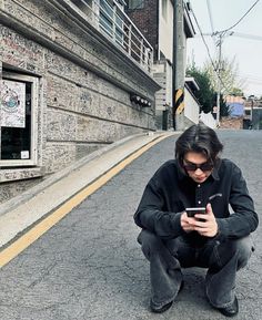 a man squatting down while looking at his cell phone on the street in front of an alleyway