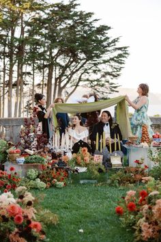 a group of people sitting in the grass with flowers around them and an open tent