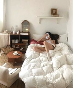 a woman laying on top of a white bed in a bedroom next to a table