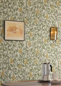 a wooden table topped with a coffee pot next to a wallpaper covered in leaves