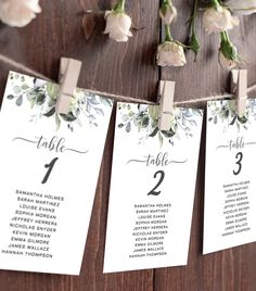 wedding seating cards hanging from clothes pins on a wooden wall with white flowers and greenery