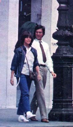 two people are walking down the sidewalk in front of a building with a clock on it