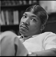 a black and white photo of a man with a tie on sitting in front of a bookshelf