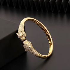 This Bracelet Is A Luxurious Gold Bangle Featuring Two Intricately Designed Panther Heads Facing Each Other At The Center. The Panthers Are Meticulously Detailed, With Sparkling Stones Encrusted Across Their Bodies, Adding A Touch Of Elegance And Brilliance To The Piece. Their Eyes Are Accentuated, Possibly With Colored Stones Or Gems, Which Adds Depth To The Animal Motif. The Bracelet Has A Sleek, Polished Gold Finish, And The Hinged Clasp Ensures A Seamless, Secure Fit. The Combination Of The Powerful Panther Design And The Dazzling Stones Make This A Statement Accessory, Symbolizing Strength, Grace, And Sophistication. This Type Of Design Is Often Favored For Its Bold Yet Timeless Aesth Panther Design, Anthropologie Jewelry, Colored Stones, Gold Bangle, High End Fashion, Gold Bangles, Bangle Bracelet, Womens Jewelry Bracelets, Gold Finish