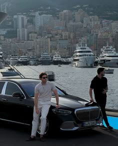 two men standing next to a black car in front of a large body of water