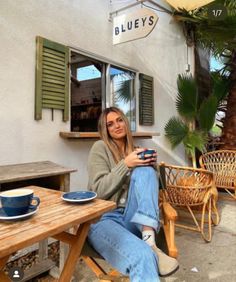 a woman sitting at a table in front of a building holding a coffee cup and looking into the camera