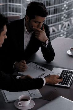 two men sitting at a table with laptops and papers