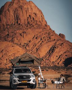 two people sitting under an umbrella next to a car in front of a large mountain