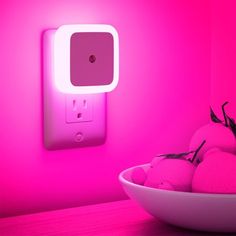 a white bowl filled with fruit next to a pink light on a wall mounted outlet