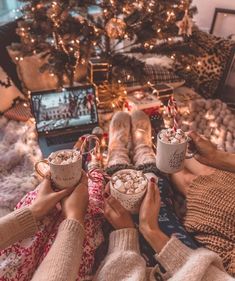 two people holding mugs with hot chocolate in front of a christmas tree