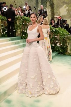 a model walks the runway in a white gown with flowers on it, while photographers take pictures behind her