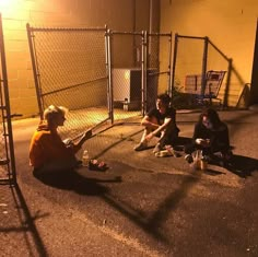 three people sitting on the ground in front of a chain link fence at night time