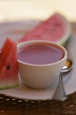 a white plate topped with a bowl of soup next to slices of watermelon