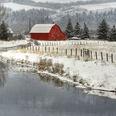 a red barn sits on the side of a snow covered hill next to a river