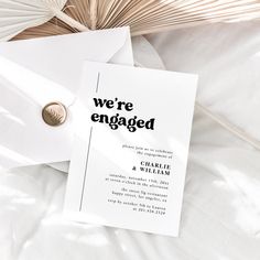 we're engaged card and envelope on top of white sheets with palm leaves in the background