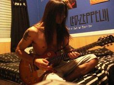 a shirtless man sitting on a bed playing an electric guitar