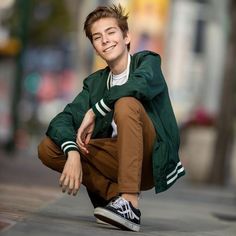 a young man crouching down on the sidewalk with his hands in his pockets and smiling