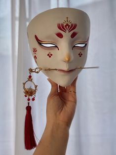 a person holding up a white mask with red and gold designs on it's face