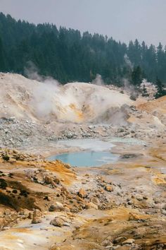 steam rises from the ground near a pool of water in a rocky area surrounded by pine trees