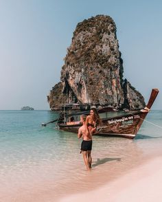 a man walking on the beach next to an old boat in the water with two large rocks sticking out of it