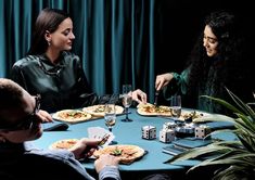 three people sitting at a table eating pizza and playing dominos on the board game