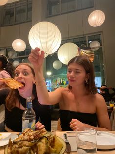 two women sitting at a table with food in front of them and one woman holding up a slice of pizza