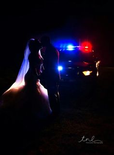 a bride and groom standing in front of a police car at night with their lights on