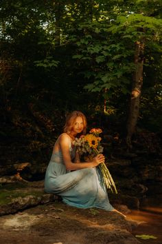 a woman sitting on the ground with flowers in her hand and trees in the background