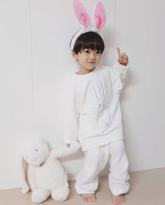 a little boy wearing bunny ears standing next to a white stuffed animal and giving the peace sign