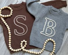 two knit sweaters with the letters s and b on them are laying next to beaded necklaces