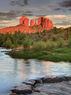 a river flowing through a lush green forest next to tall red rock formations in the distance