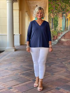 a woman in white pants and a blue top is standing on a brick walkway with her hands in her pockets