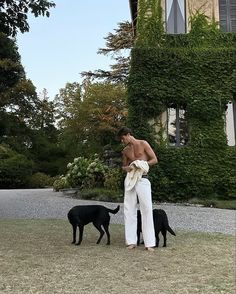 a shirtless man standing next to two black dogs in front of a large house