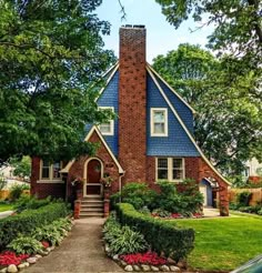 a brick house with blue shingles and red flowers in the front yard, surrounded by trees