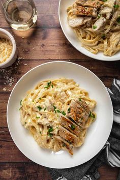 two plates of pasta with chicken and parmesan cheese on the side next to a glass of wine