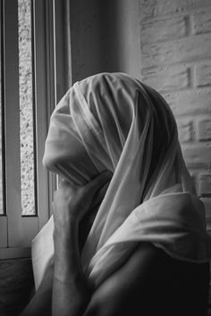 black and white photograph of a woman with her head covered by a blanket looking out the window