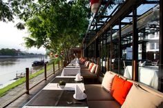 an outdoor dining area with tables and chairs on the side of a river bank, along with large glass windows