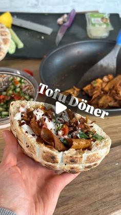 a person holding up a sandwich in front of some other food on a wooden table