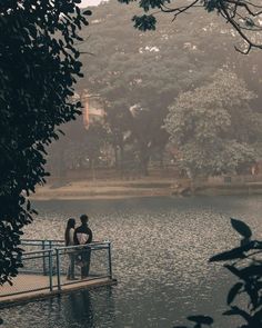 two people standing on a pier over a body of water with trees in the background