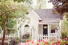 a small white house with flowers in the front yard and on the side, there is a picket fence