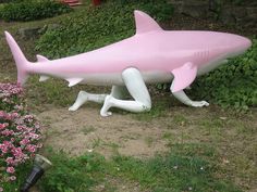 a pink and white fake shark sitting on top of a lush green field next to flowers